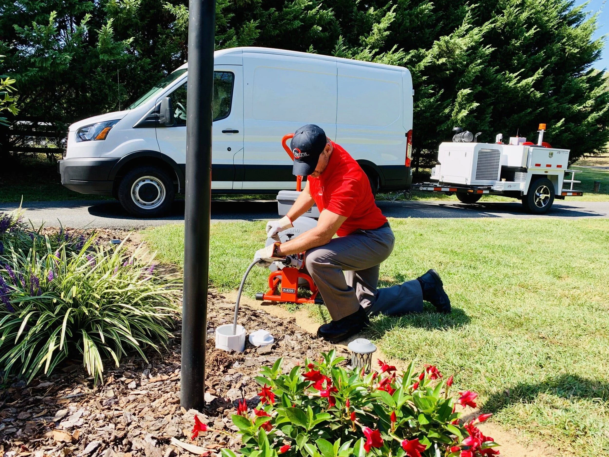 A worker kneeling on the grass, using equipment near a pole and a garden bed with plants and red flowers, with a white utility van and a trailer in the background on a sunny day.