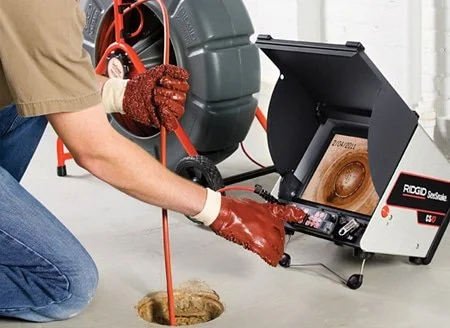 A person wearing red gloves kneels beside a drain and uses a pipe inspection camera connected to a portable monitor to inspect the drain, with a portable incinerator nearby.