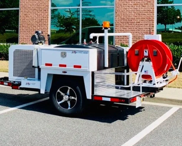 A portable fire pump trailer with a red hose reel, mounted on a white metal frame with a black water tank and engine, parked in a parking lot outside a brick building.
