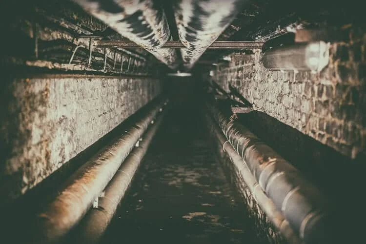 A view down a narrow, dark underground tunnel with brick walls and metal pipes running along each side.