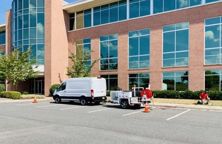 A white service van parked in front of a modern office building with large windows, with a work trailer and orange cones nearby, and a worker in red attending to equipment.
