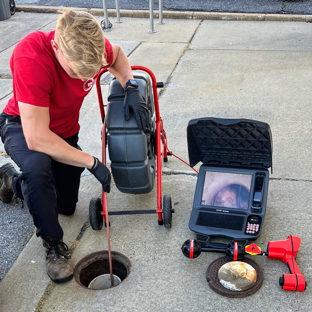 A person wearing a red shirt and black gloves kneels on the sidewalk, inspecting a sewer access point with a robotic camera device. The equipment includes a portable control unit with a screen and a robotic crawler with a camera attached, used for sewer inspection.