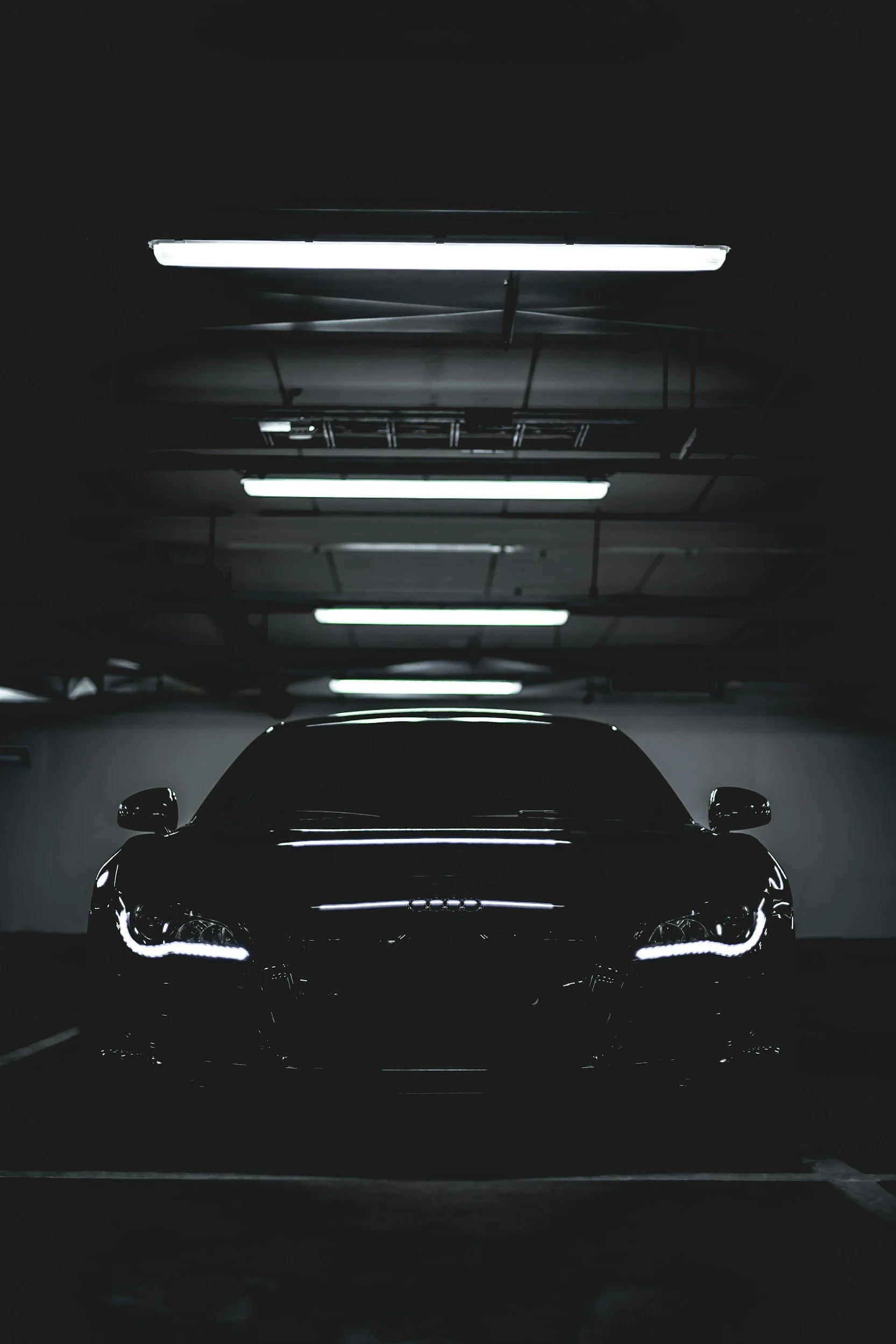 A black Audi car parked in a dimly lit underground garage with fluorescent ceiling lights.