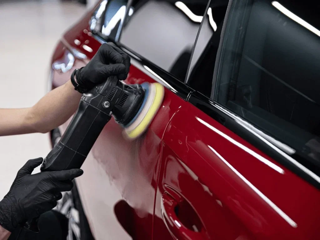 Person polishing a red car with a buffer machine, wearing black gloves.