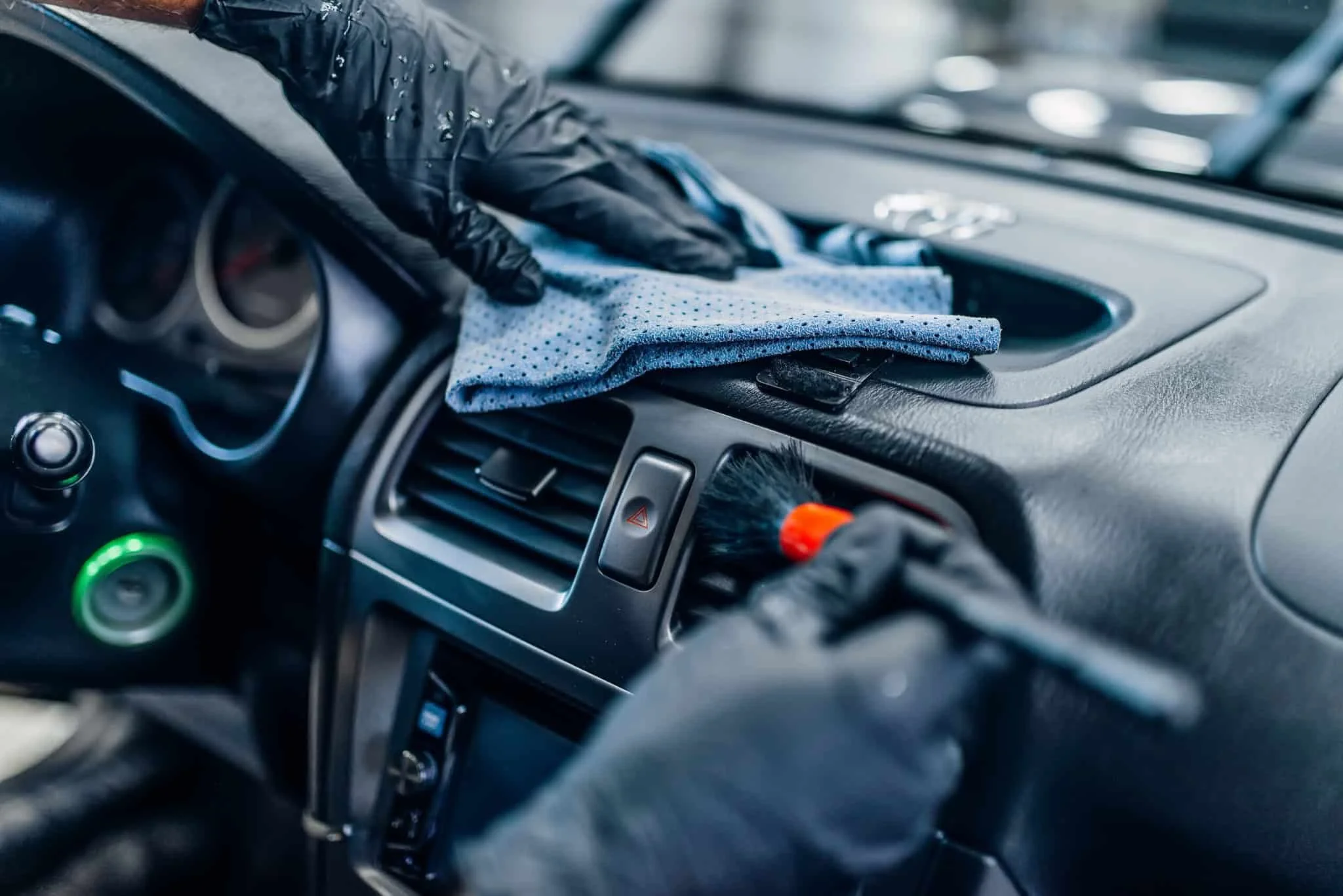 Person cleaning the dashboard of a car with a cloth and a duster, wearing black gloves.