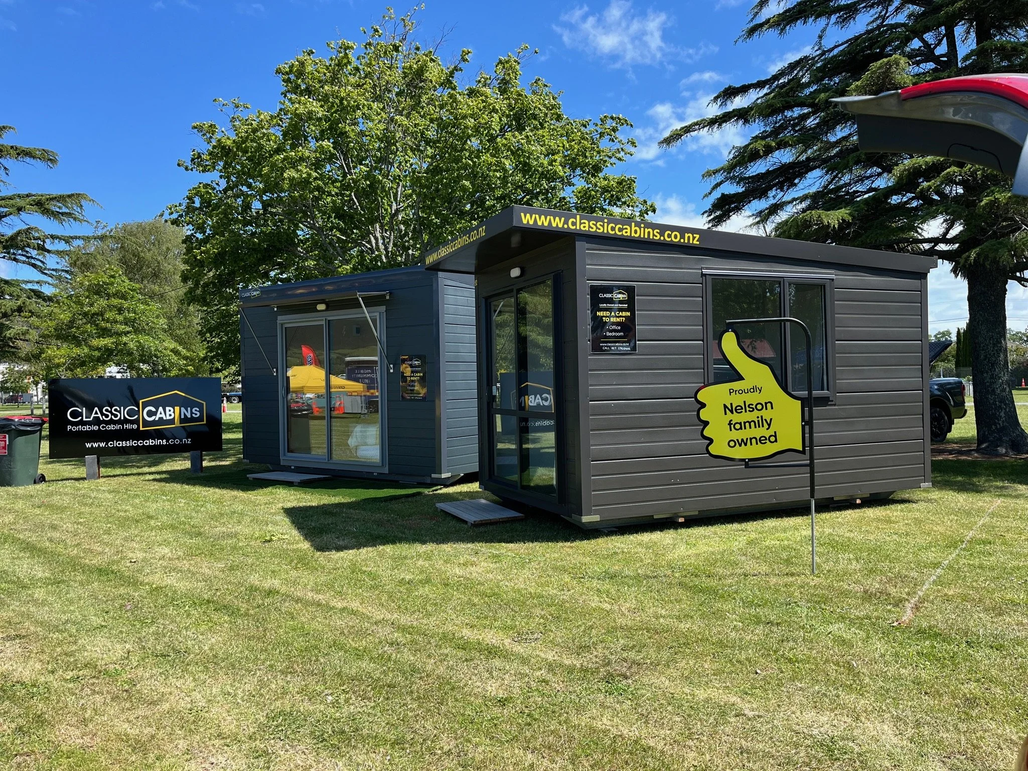 Display of two compact, dark gray portable cabins with large windows, situated on a grassy area with trees in the background and a blue sky. A sign beside them advertises classic cabins for rent, and a yellow sign states the Nelson family owns the cabins.