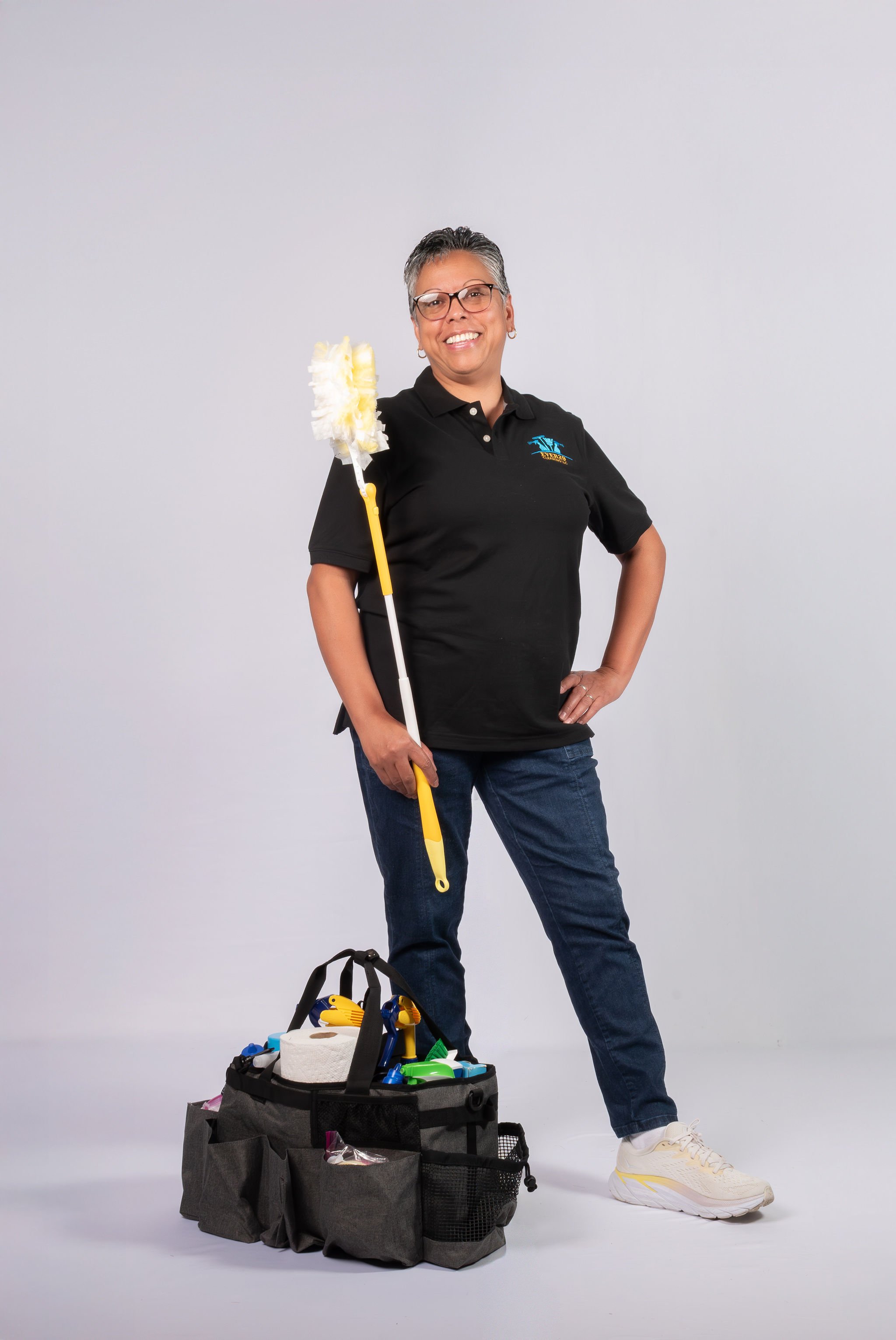 A woman with short gray hair, glasses, and wearing a black polo shirt and jeans, standing with one hand on her hip, holding a cleaning duster in the other, next to a black cleaning supply bag filled with cleaning tools, against a plain gray background.