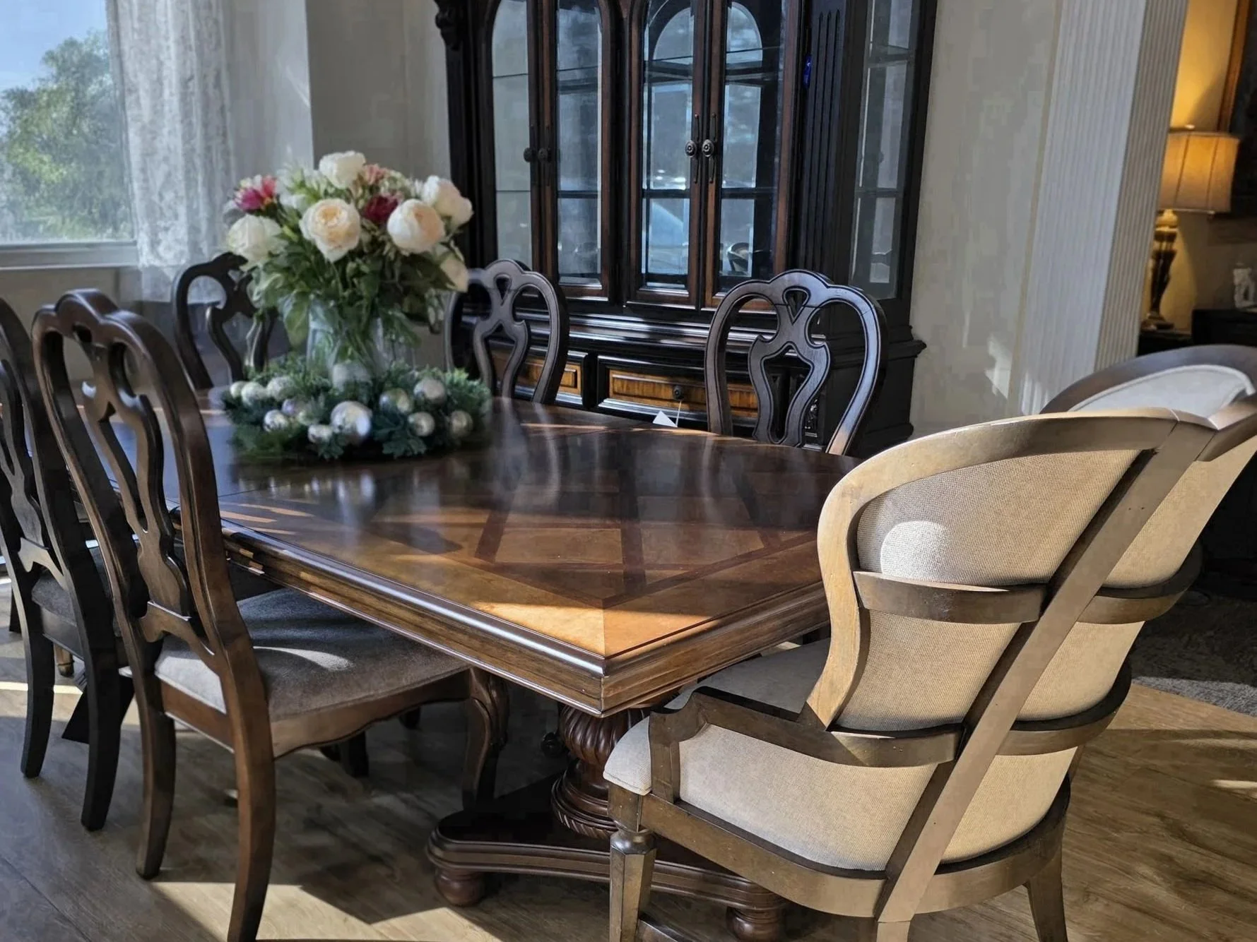 Dining room with a wooden table, floral centerpiece with pink and white flowers, chairs surrounding the table, a window with sheer curtains, and a china cabinet in the background.
