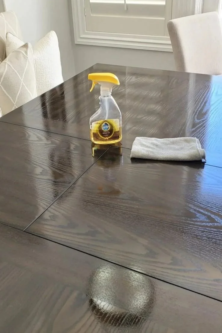 A wooden dining table with a spray bottle of cleaning solution and a folded cloth on it, with white cushions and a sliding window in the background.