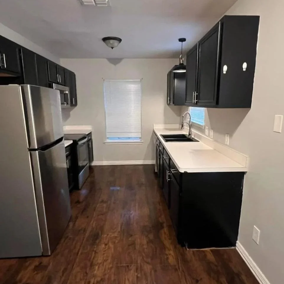 Empty kitchen with black cabinets, stainless steel refrigerator, black stove, white countertops, hardwood flooring, and a small window with blinds.