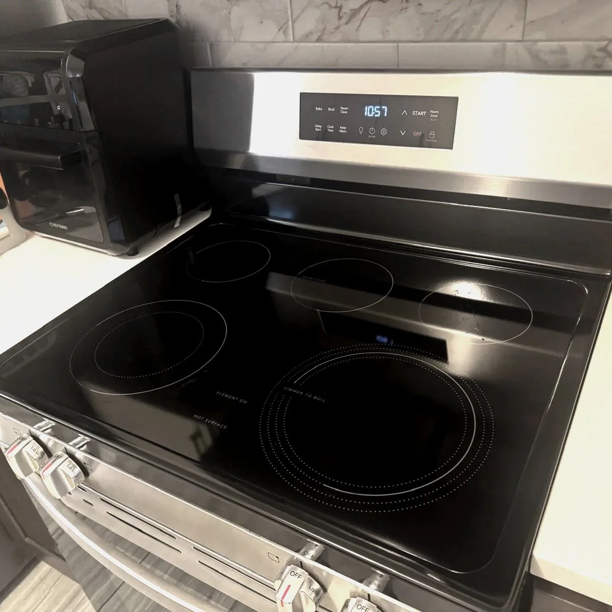 Stainless steel electric stove with four burners and digital clock displaying 10:57, located in a kitchen with gray tile backsplash and a black microwave oven on the counter nearby.