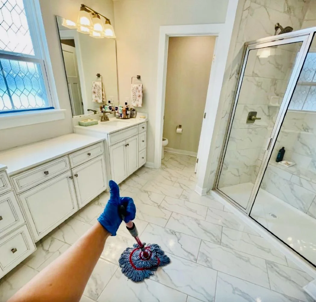 A person wearing a blue glove is mopping a marble floor in a bright bathroom with a white vanity, a large mirror, a window, and a glass shower stall.