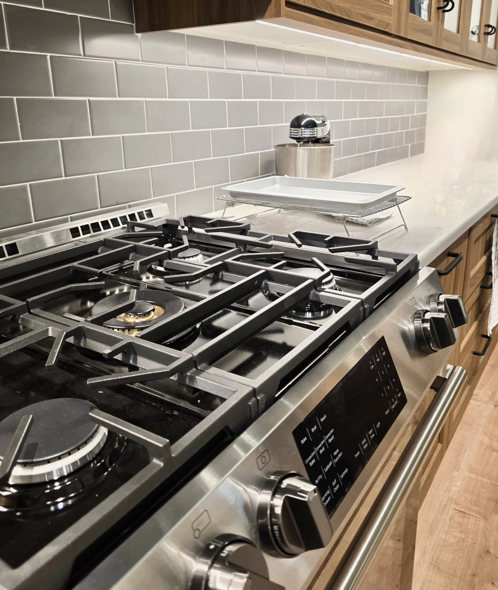 A kitchen stove with a gray tile backsplash and a white quartz countertop. There are stacked white dishes on a metal rack and a small stainless steel appliance on the countertop in the background.