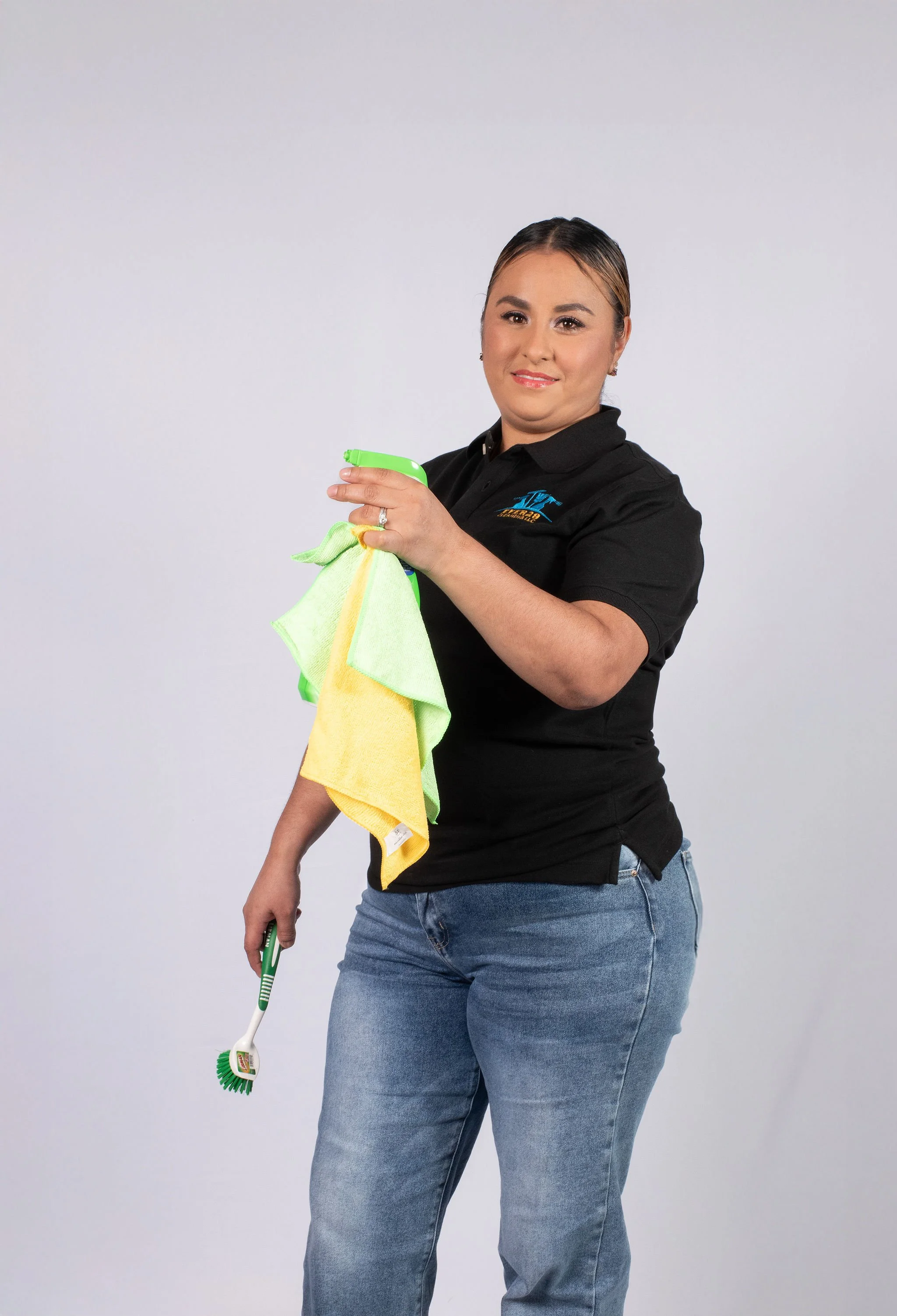 A woman dressed in a black polo shirt and blue jeans holding cleaning supplies, including a yellow and green cloth and a scrub brush, standing against a plain white background.