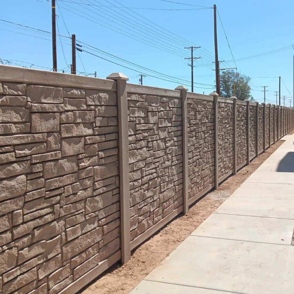 A long stone-patterned fence with evenly spaced vertical posts runs alongside a sidewalk under a clear blue sky with power lines overhead.