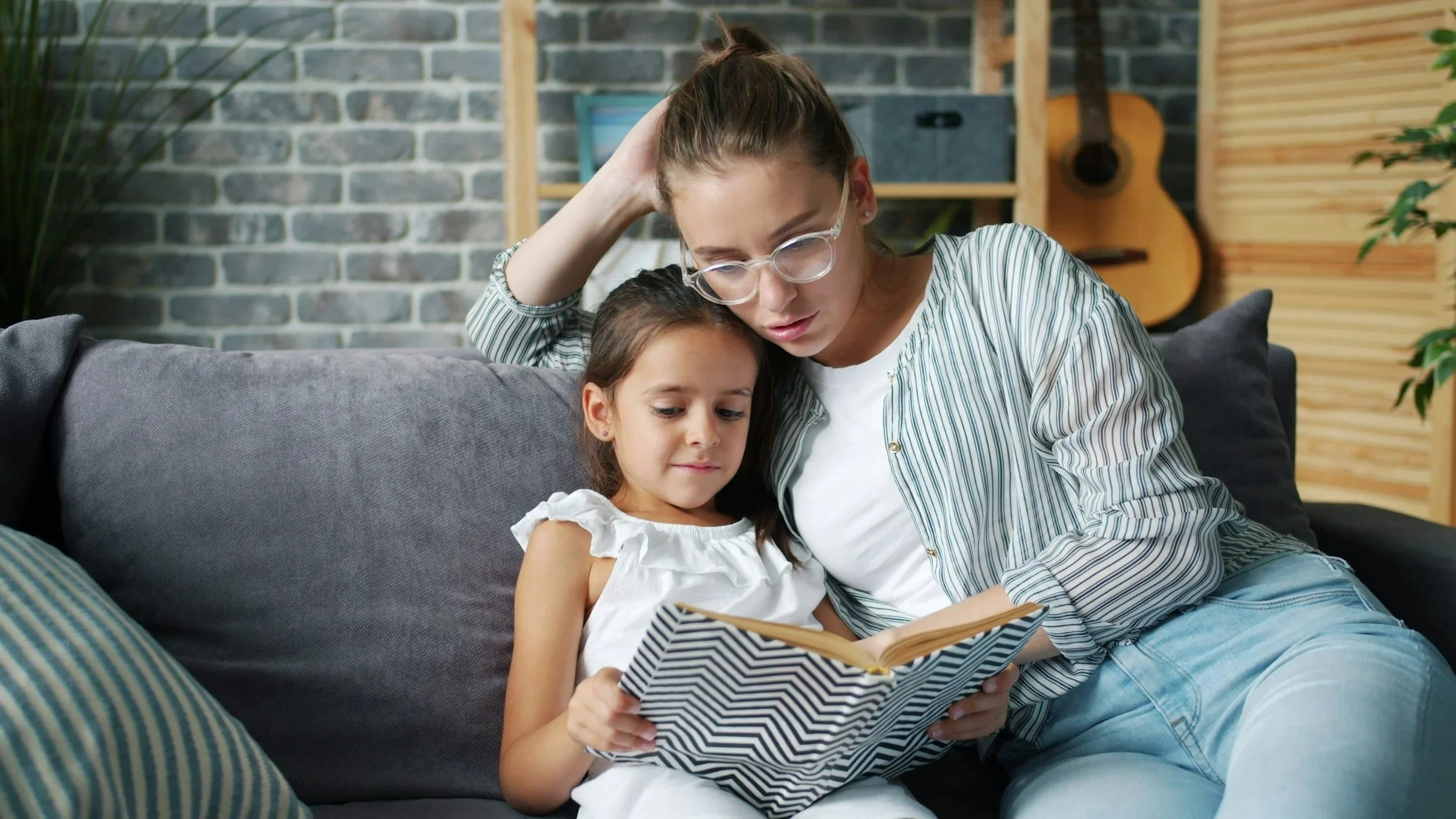 A woman and a young girl sit on a couch looking at a book together in a living room with a brick wall, a guitar hanging on the wall, and a plant in the background.