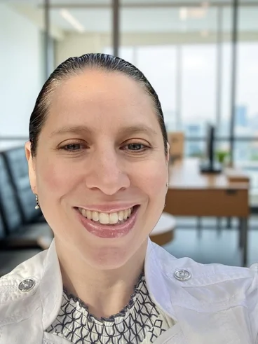 A woman takes a selfie in a modern office with large windows and a desk in the background.