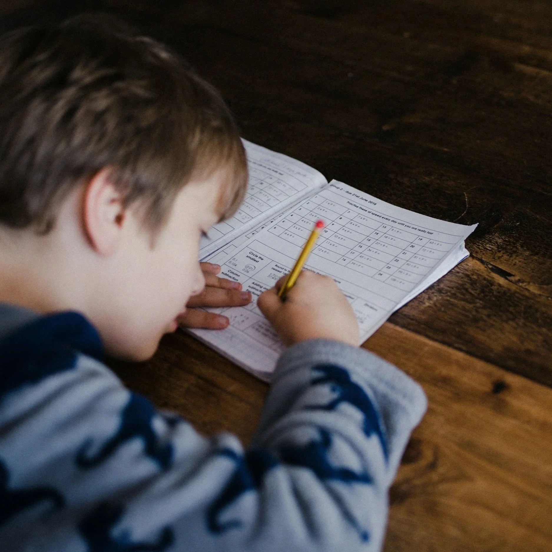A young boy with light brown hair, wearing a gray and navy blue hoodie, is lying on a wooden table while doing homework with a pencil in his hand.