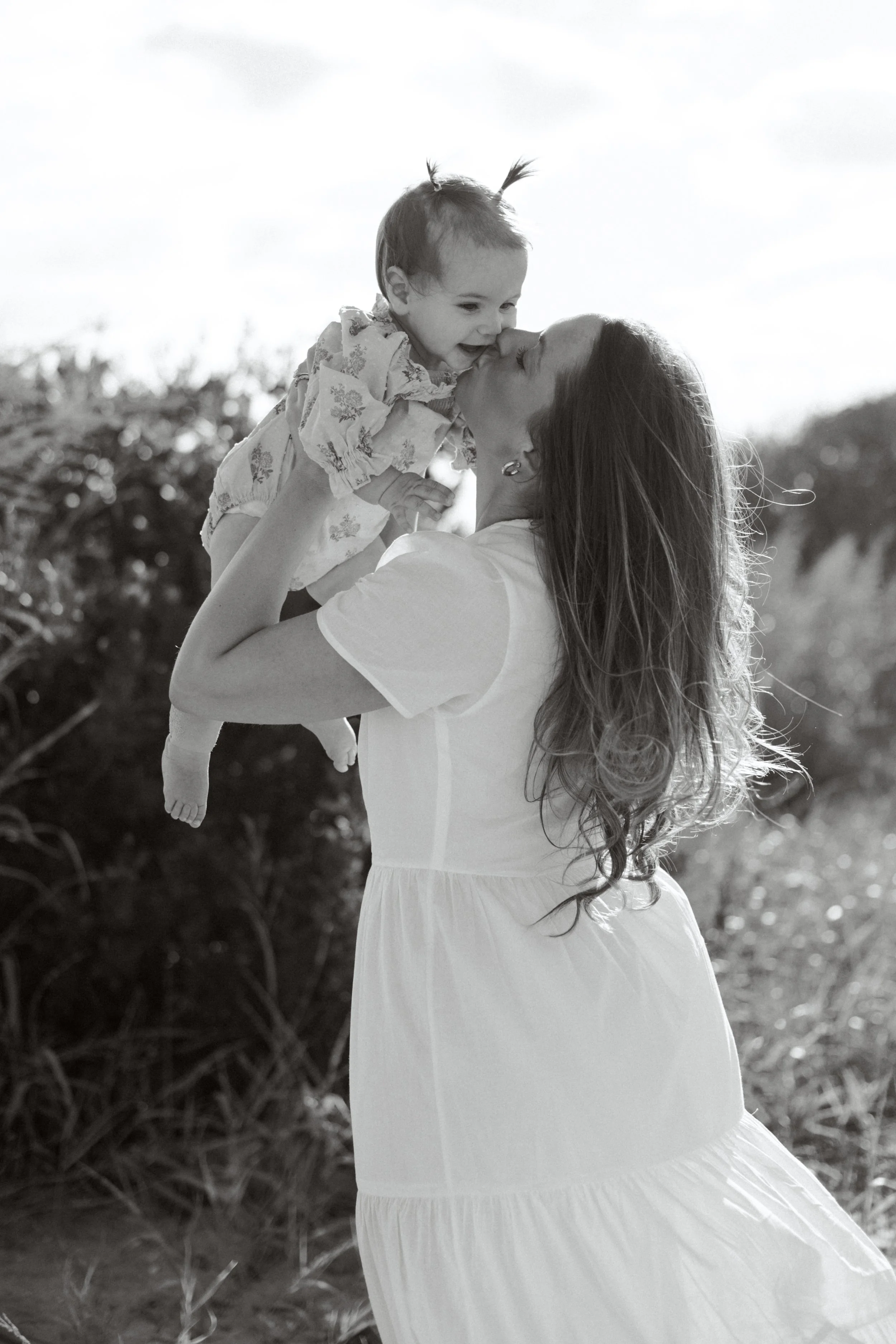 A woman holding a baby girl in an outdoor setting during the daytime. The woman is lifting the baby up, and the baby is touching her face with her hands. The scene is bright and cheerful with natural background.