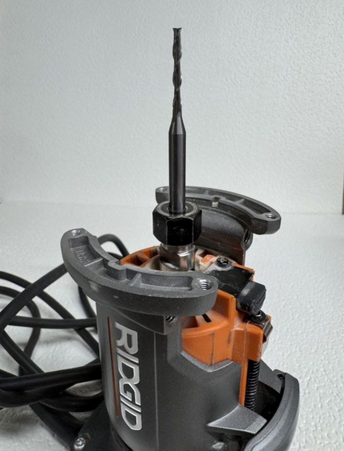 Close-up of a router with a drill bit attached, placed on a gray surface against a white wall.