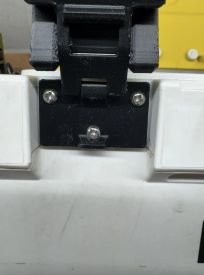 Close-up of a black mounting bracket attached to a white surface with screws, in front of a yellow object.