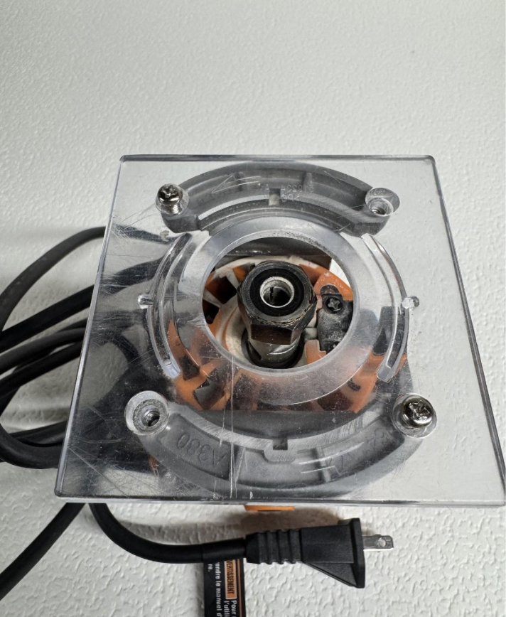 Close-up of a router with a clear plastic safety cover, showing the internal collet and bit; power cord attached.