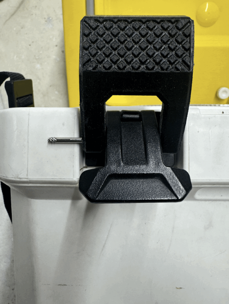 A black adjustable foot pedal on a white surface, with a textured grip on top, attached to a gray cord. There is a yellow and white object in the background.