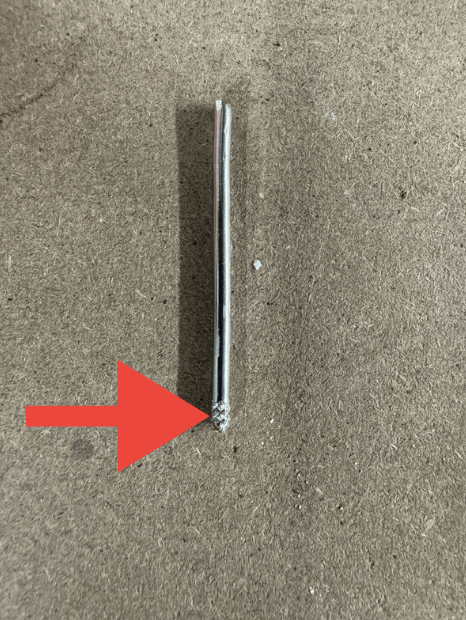 Close-up of a broken metal object, possibly a nail or screw, on a brown surface with a red arrow pointing to the damaged end.