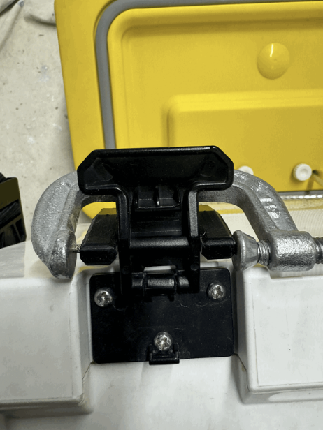 Close-up of a black latch mechanism attached to a white surface with a yellow object in the background.