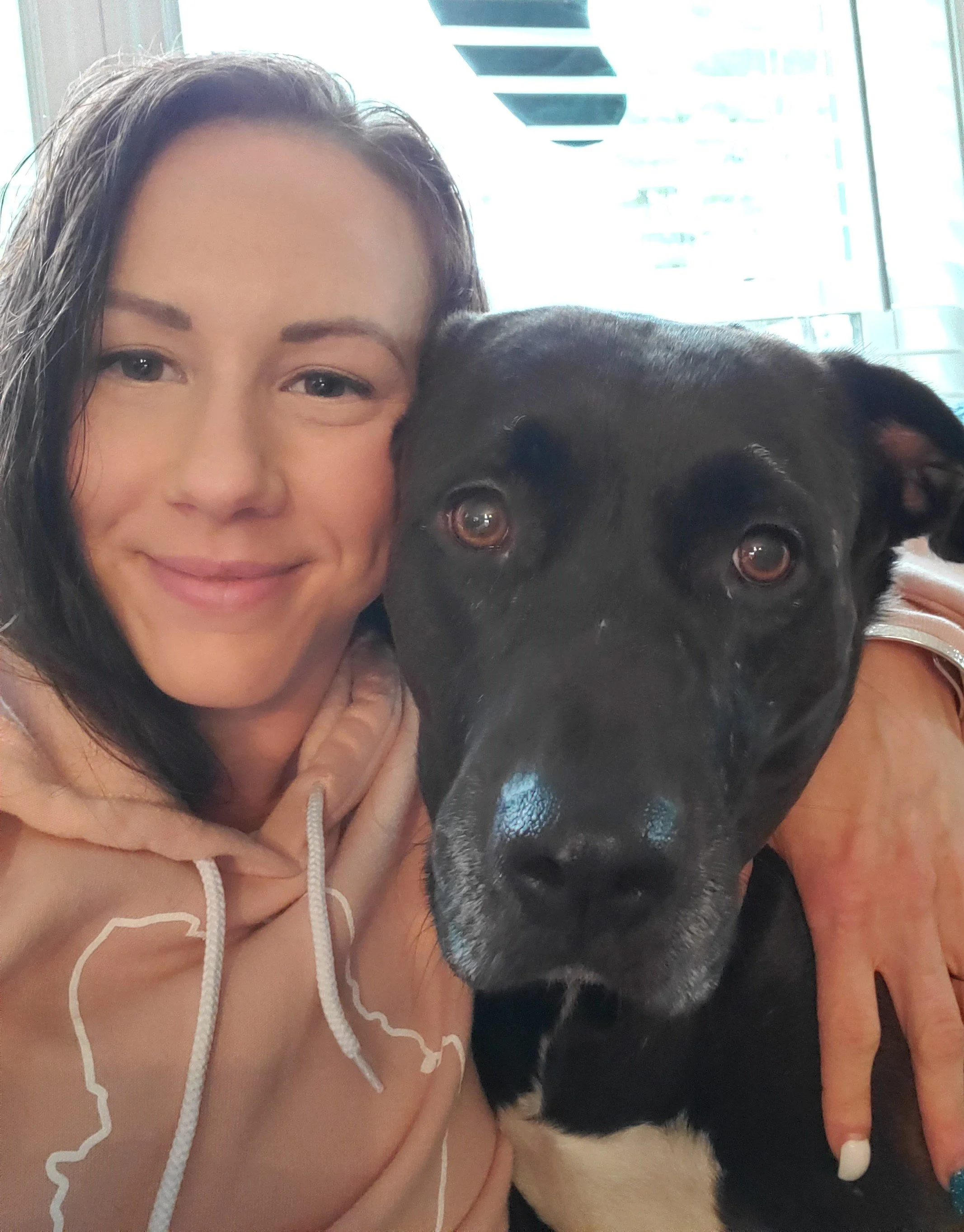 A woman with dark hair and a light brown hoodie smiling and hugging a large black dog with brown eyes inside a building with large windows and modern design elements.