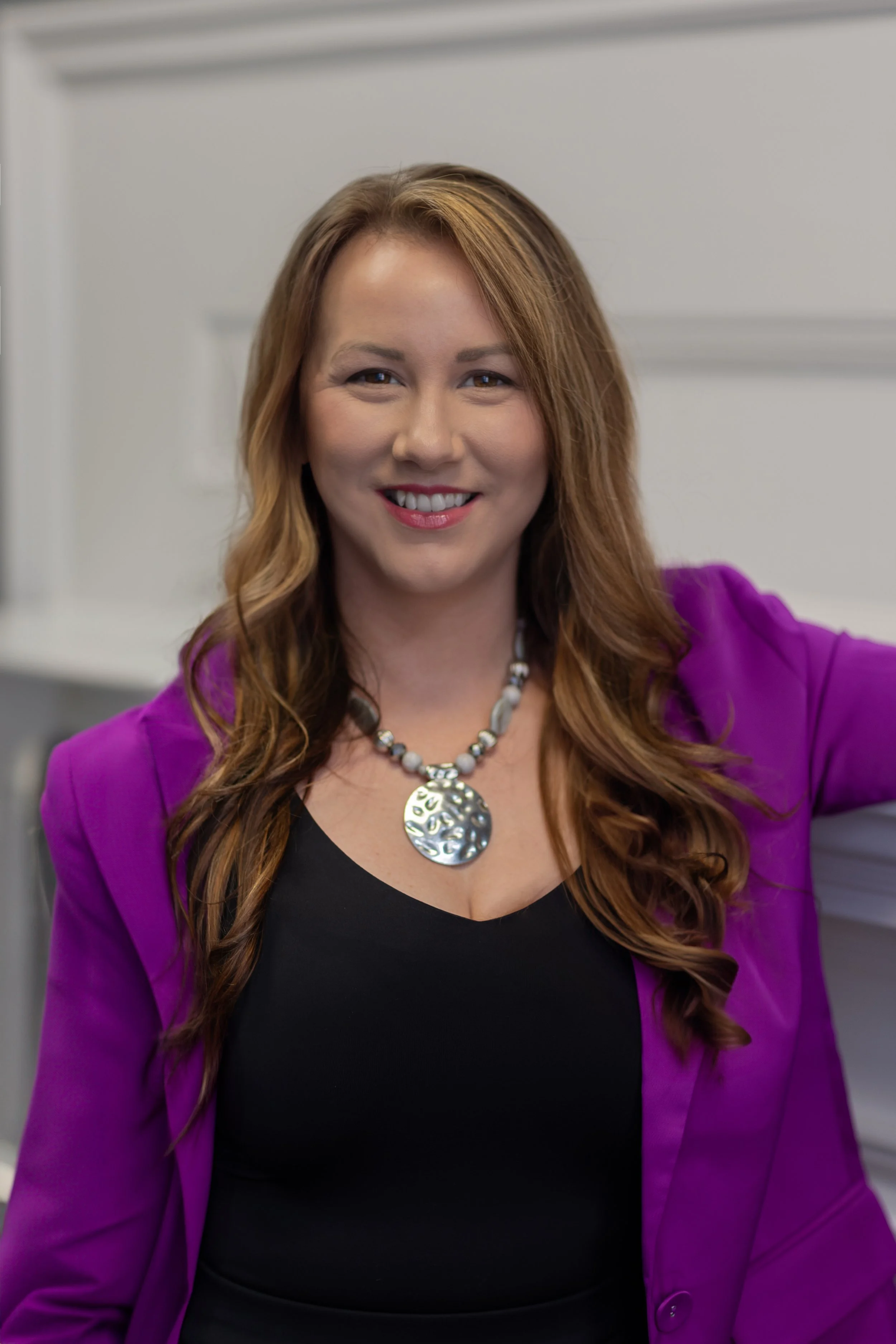A smiling woman with wavy brown hair wearing a black top, purple blazer, and a large silver necklace, leaning against a white counter or shelf in an indoor setting.