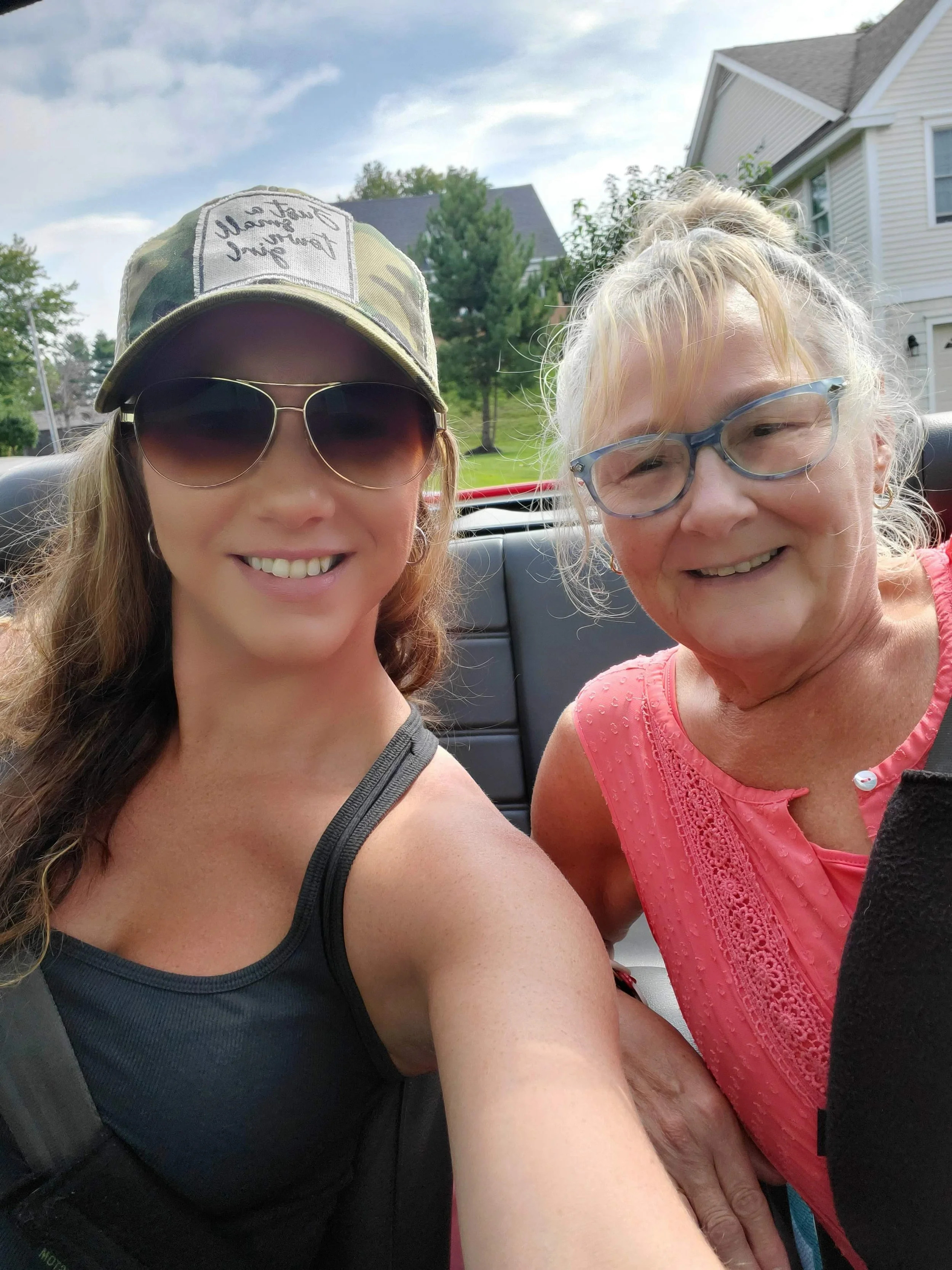 Two women smiling in a convertible car with houses and trees in the background.