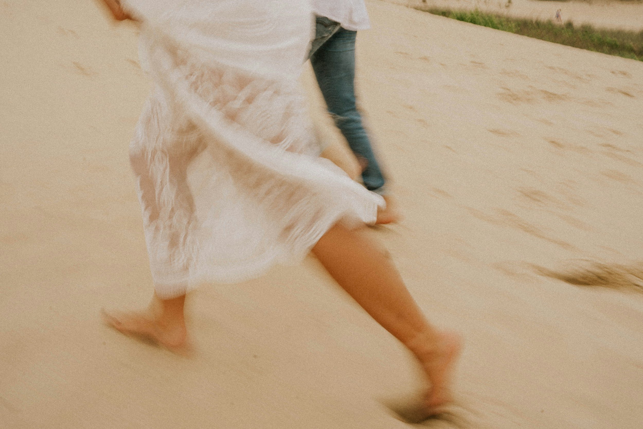 Blurred image of two people running, one wearing a white dress and the other wearing blue jeans, on a sandy beach.