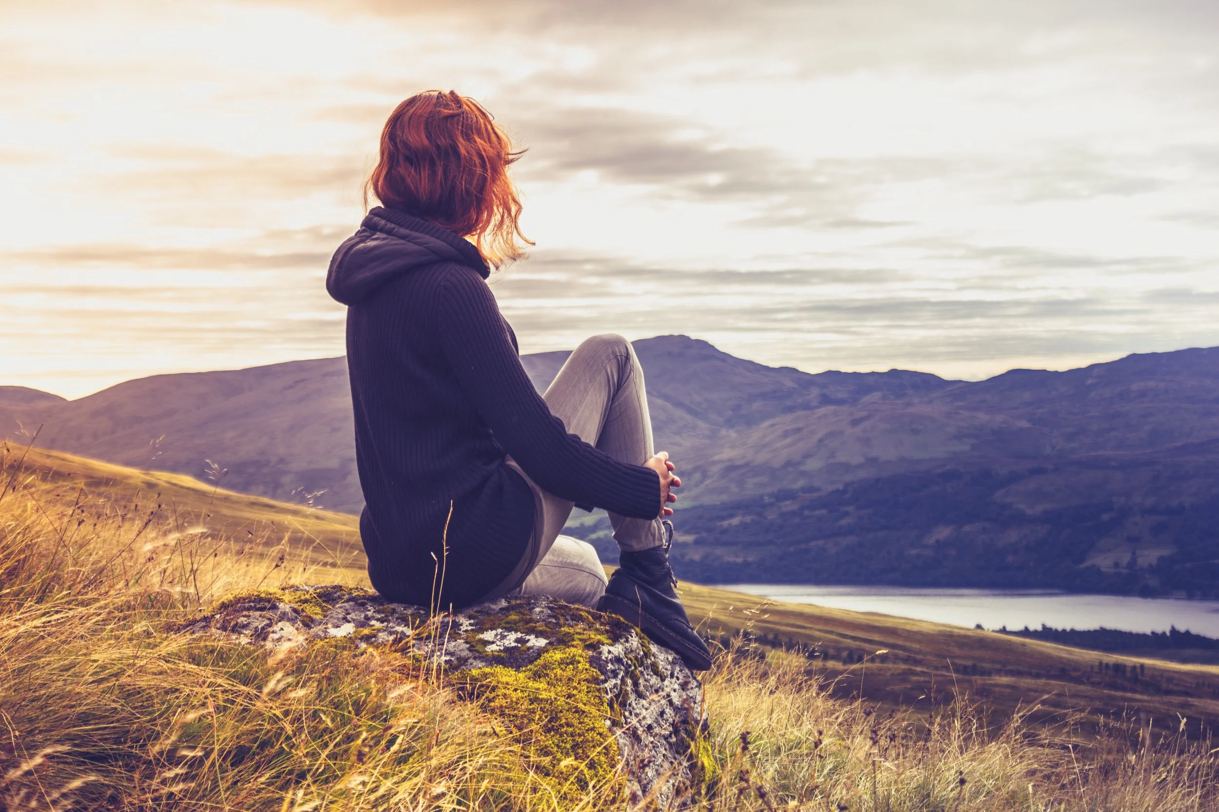 A person with red hair sitting on a rock in a grassy field, overlooking a mountain landscape and a body of water at sunset.