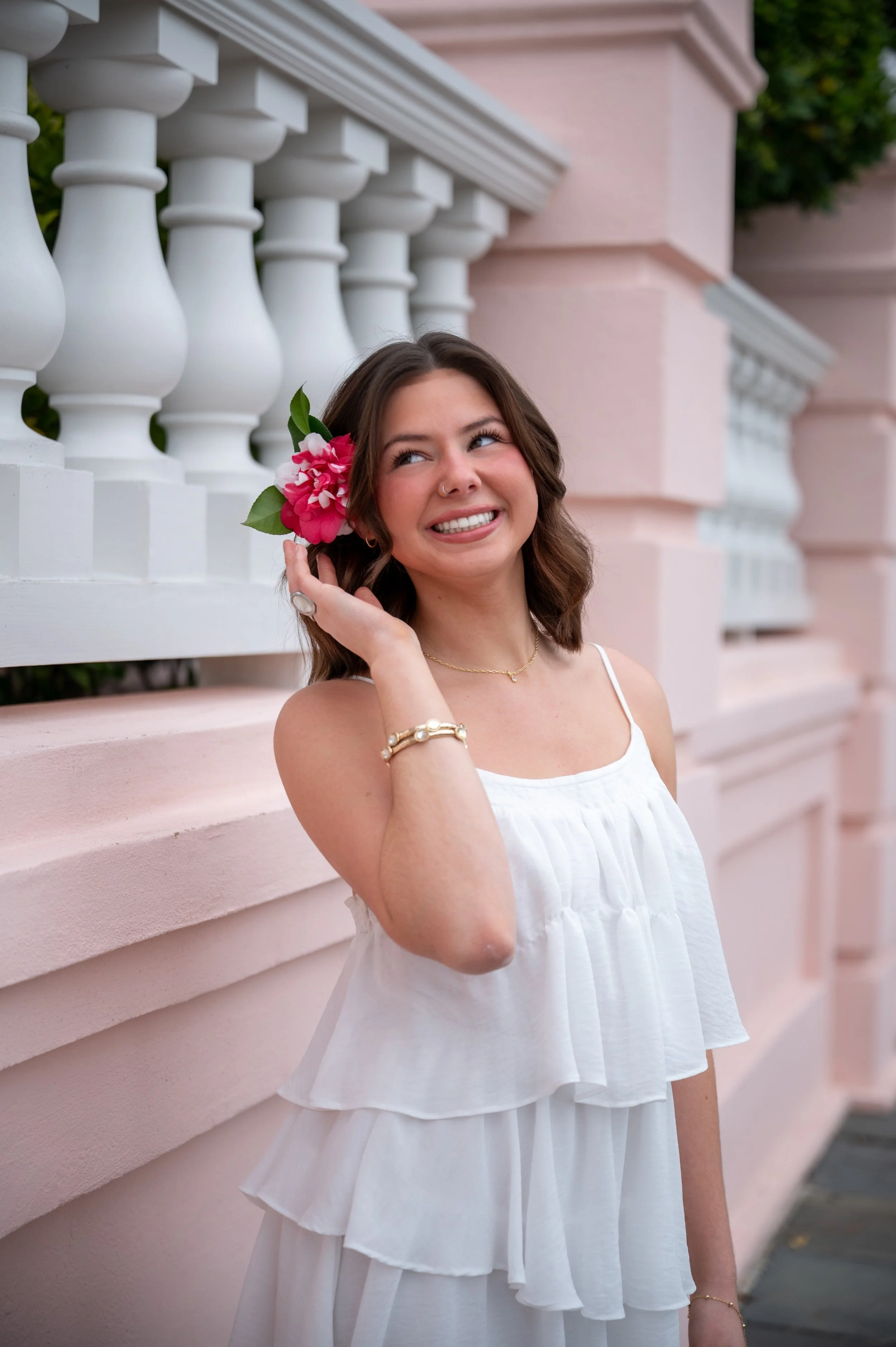 A young woman in a white sleeveless dress holds a pink flower near her ear and smiles, standing beside a pink wall with white railings.