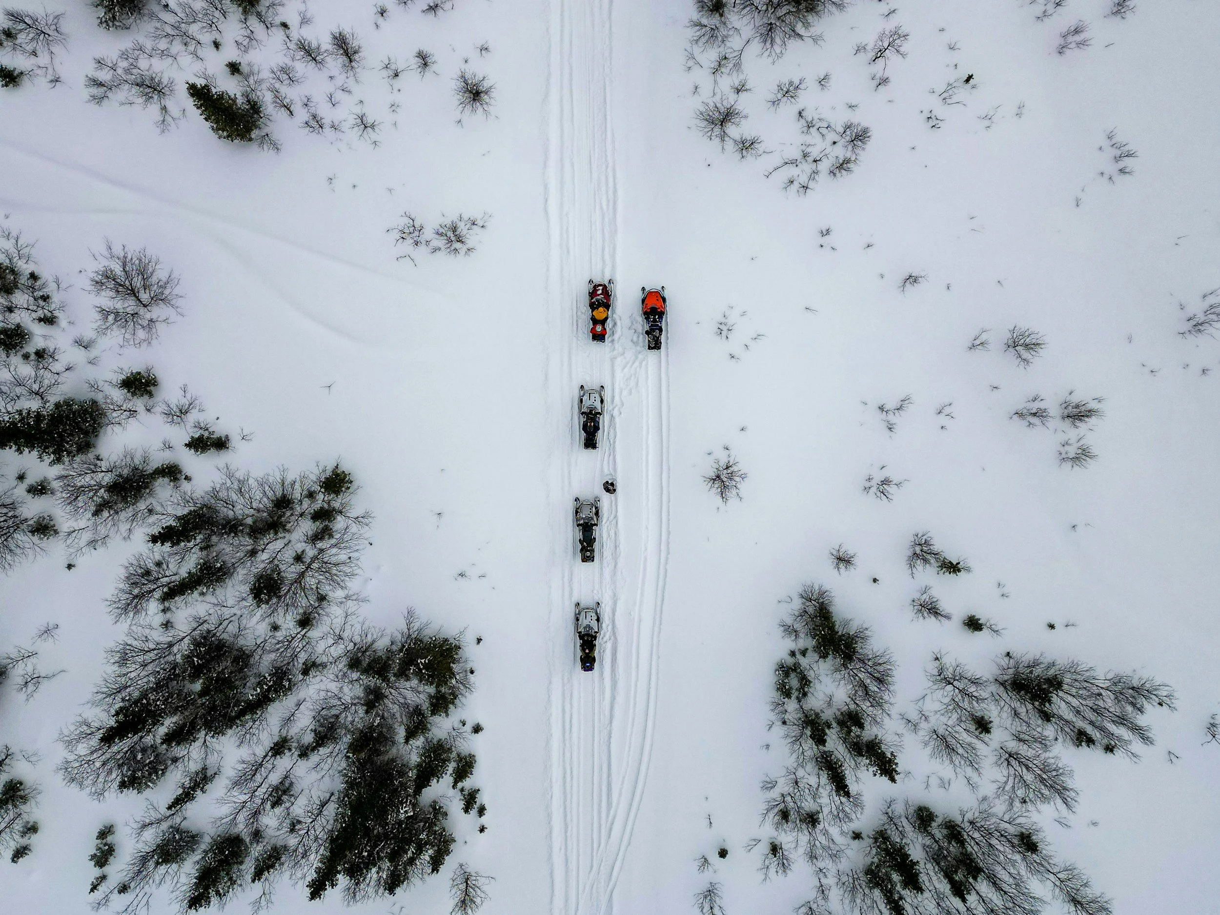 Aerial view of snow-covered landscape with trees and a snowmobile trail. Six snowmobiles are riding in a line on the trail.