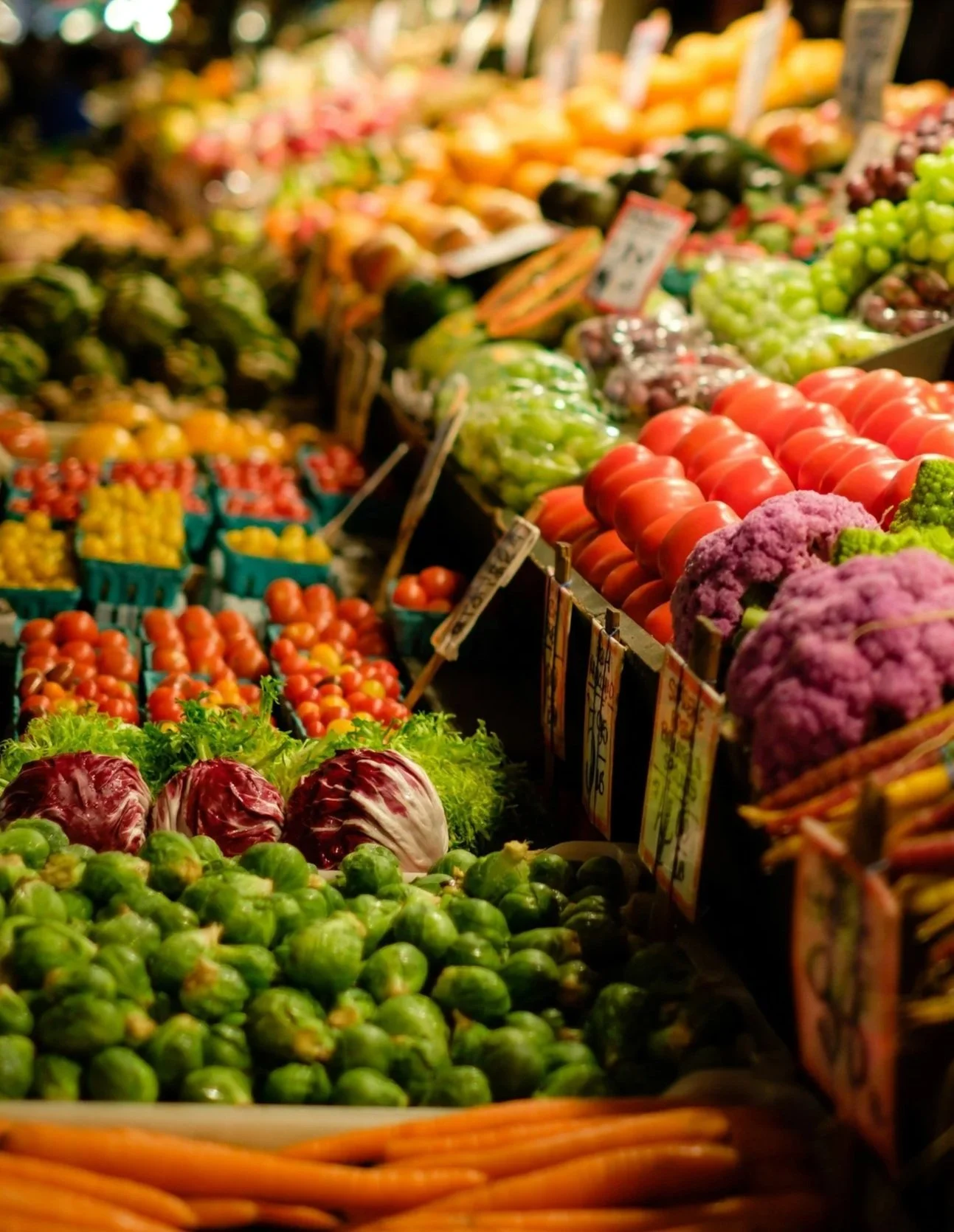 A display of fresh vegetables at a grocery store, including Brussels sprouts, radicchio, carrots, tomatoes, cauliflower, and other produce.