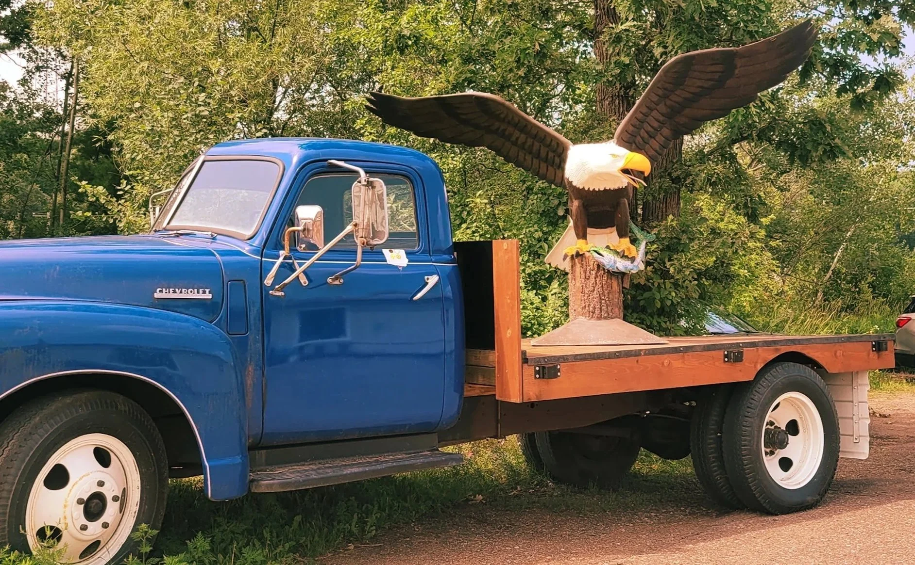 A vintage blue Chevrolet truck with a wooden flatbed, featuring a large wooden eagle sculpture with outstretched wings attached to a tree trunk on the flatbed.