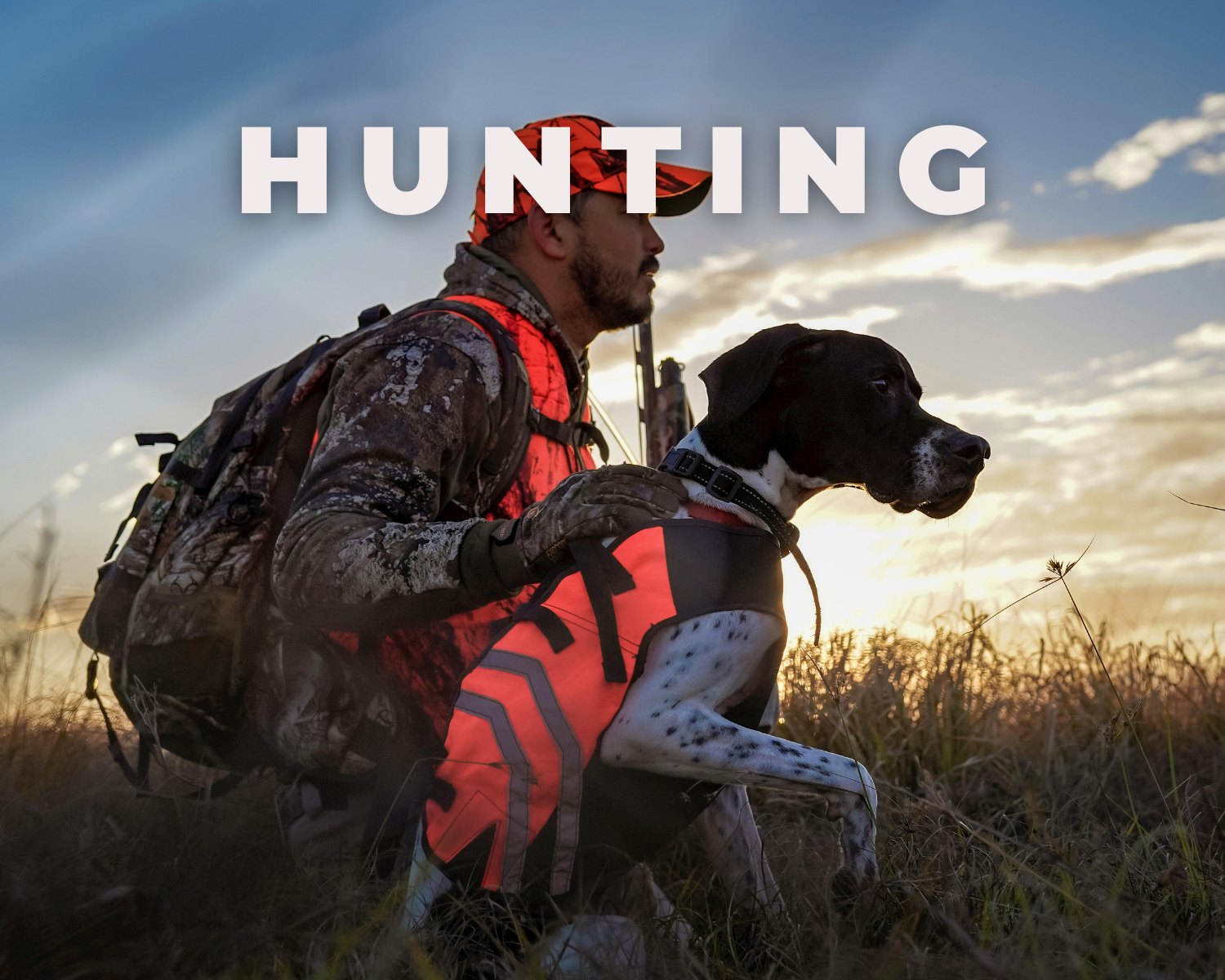 A man dressed in camouflage and orange hunting gear with a hunting dog sitting on tall grass at sunset.