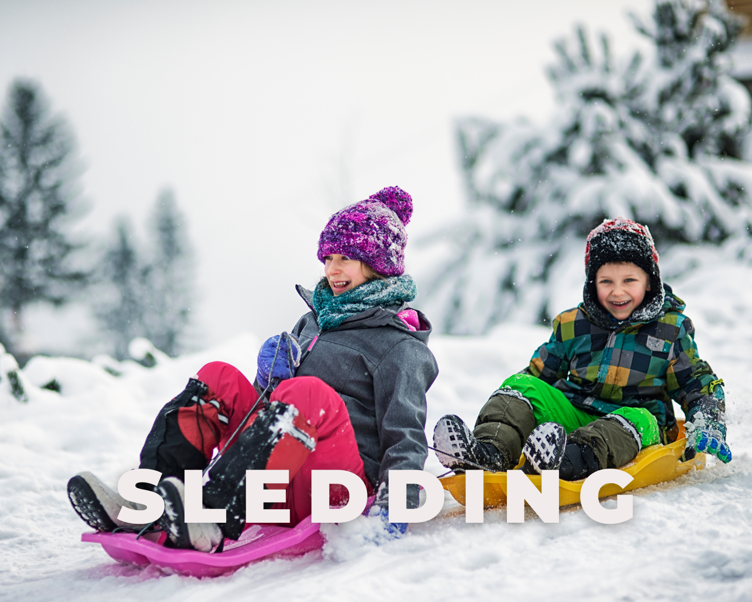 Two children sledding down a snowy hill, smiling and enjoying winter weather, with snow-covered trees in the background.