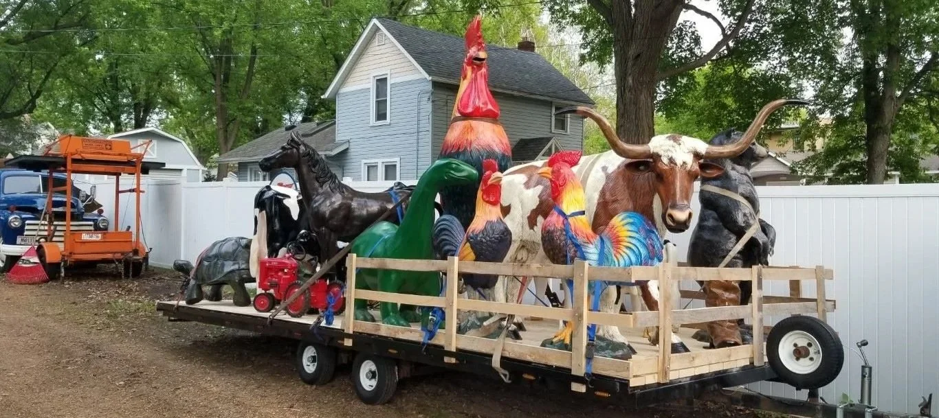 Trailer loaded with large colorful animal statues including a rooster, a horse, a rooster, a longhorn steer, a duck, and a pheasant, parked outdoors with trees and residential houses in the background.