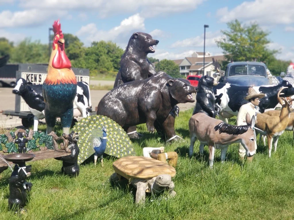 Outdoor display of animal sculptures including a rooster, bears, cows, a turtle, a peacock, a horse, and a hiker figure on grass with buildings and vehicles in the background.