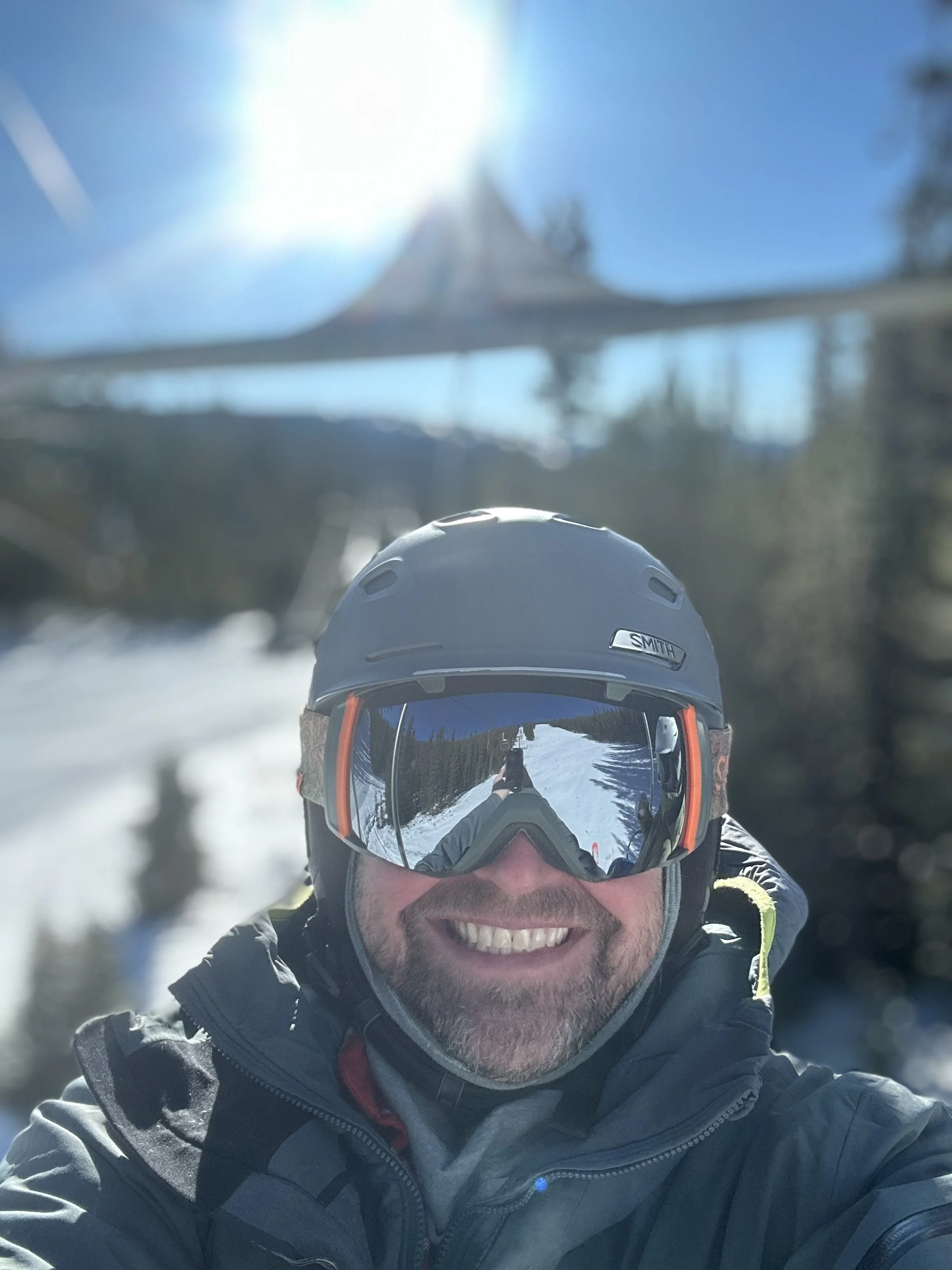 A man smiling, wearing ski gear including a helmet and reflective ski goggles, in a snowy outdoor setting with trees and a ski lift in the background.