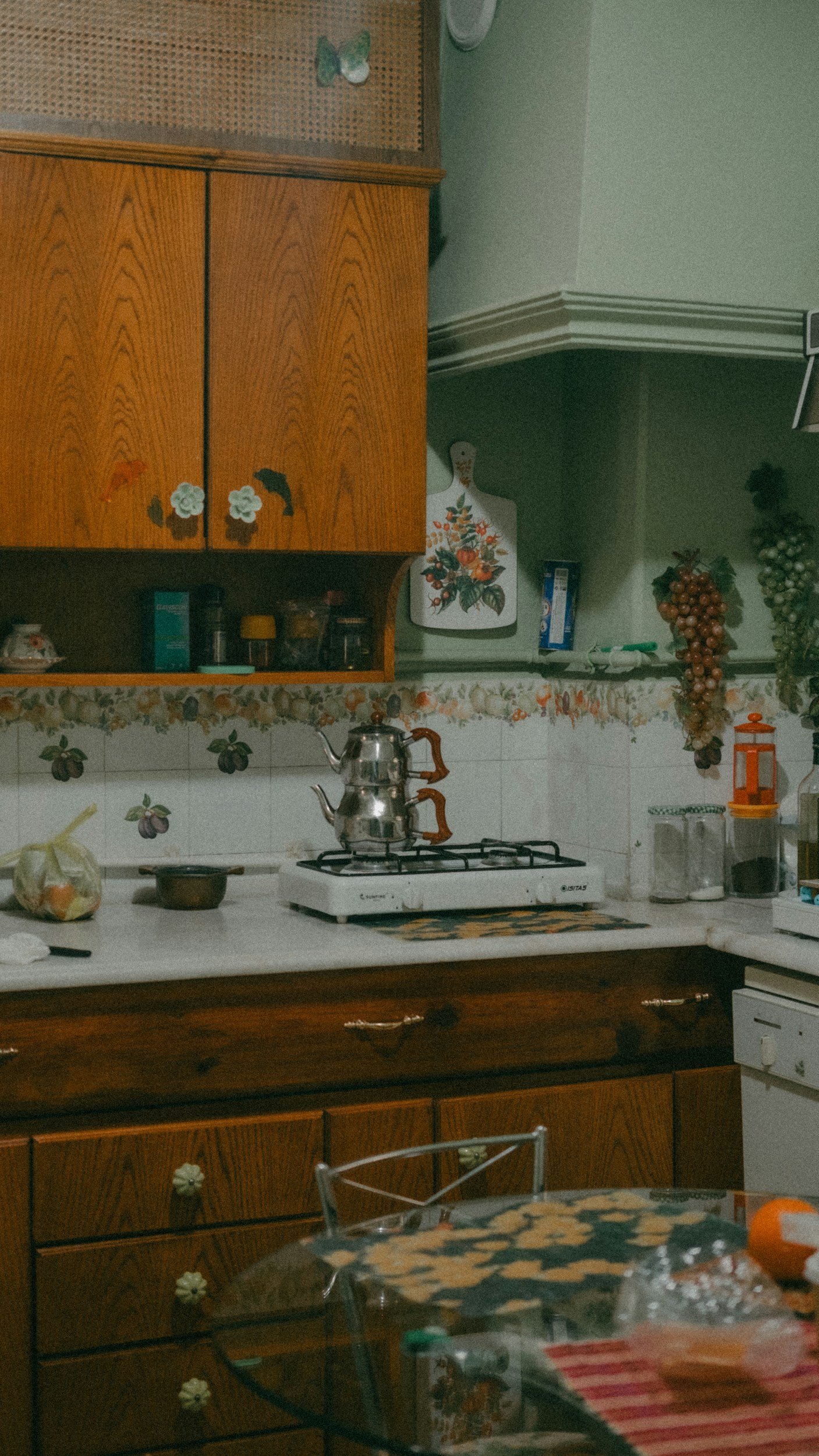 A kitchen with wooden cabinets, a gas stove, and a cluttered countertop. Decorative grapes and a cutting board with a floral design are on the wall. There are various kitchen items, including a teapot on the stove, a bag on the counter, and a glass table with items on it in the foreground.