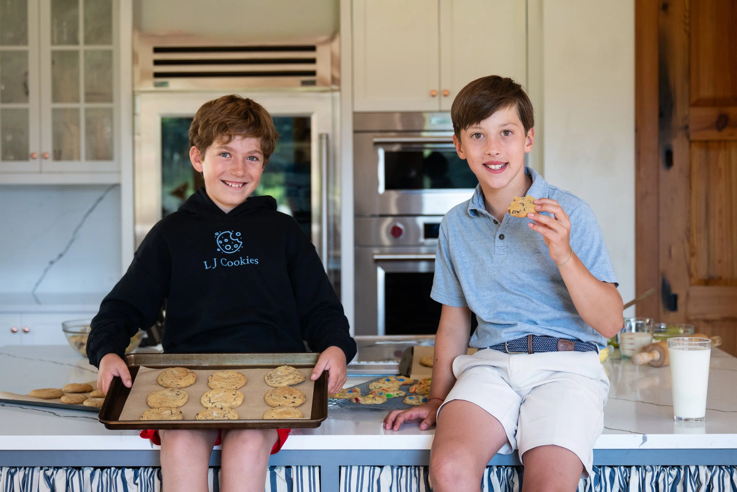 Two boys sitting on a kitchen counter, one holding a tray of freshly baked chocolate chip cookies, the other eating a cookie, with a glass of milk nearby.