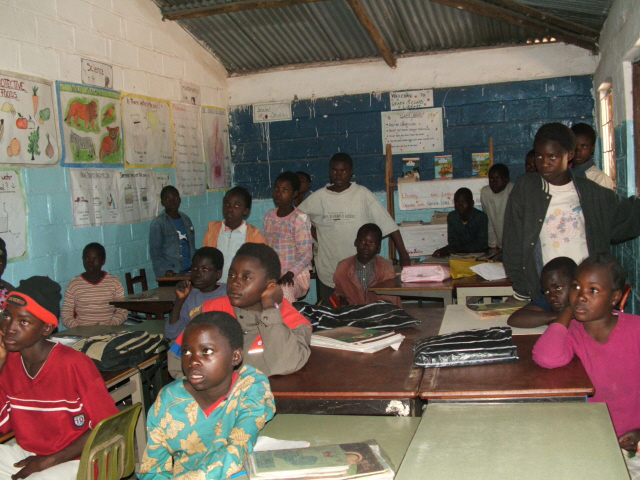 School classroom with young students paying attention, some seated and some standing by desks, with educational posters on the wall.