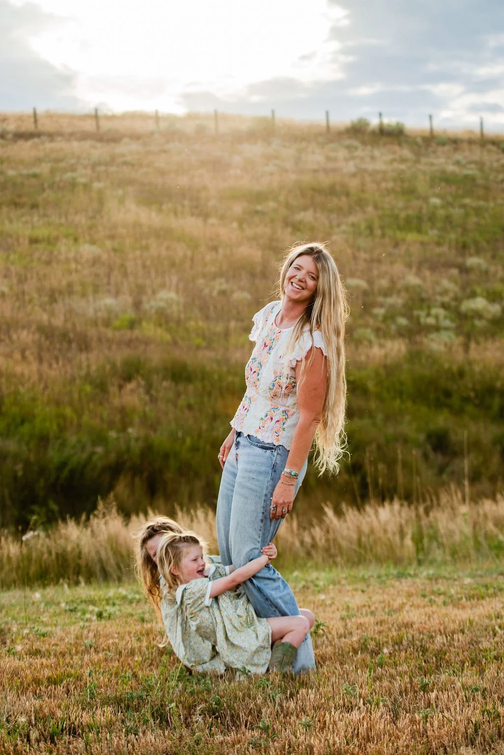 A woman with long blonde hair standing in a grassy field with two young girls, one hugging her leg and the other sitting on the ground, all smiling happily outdoors during the daytime.