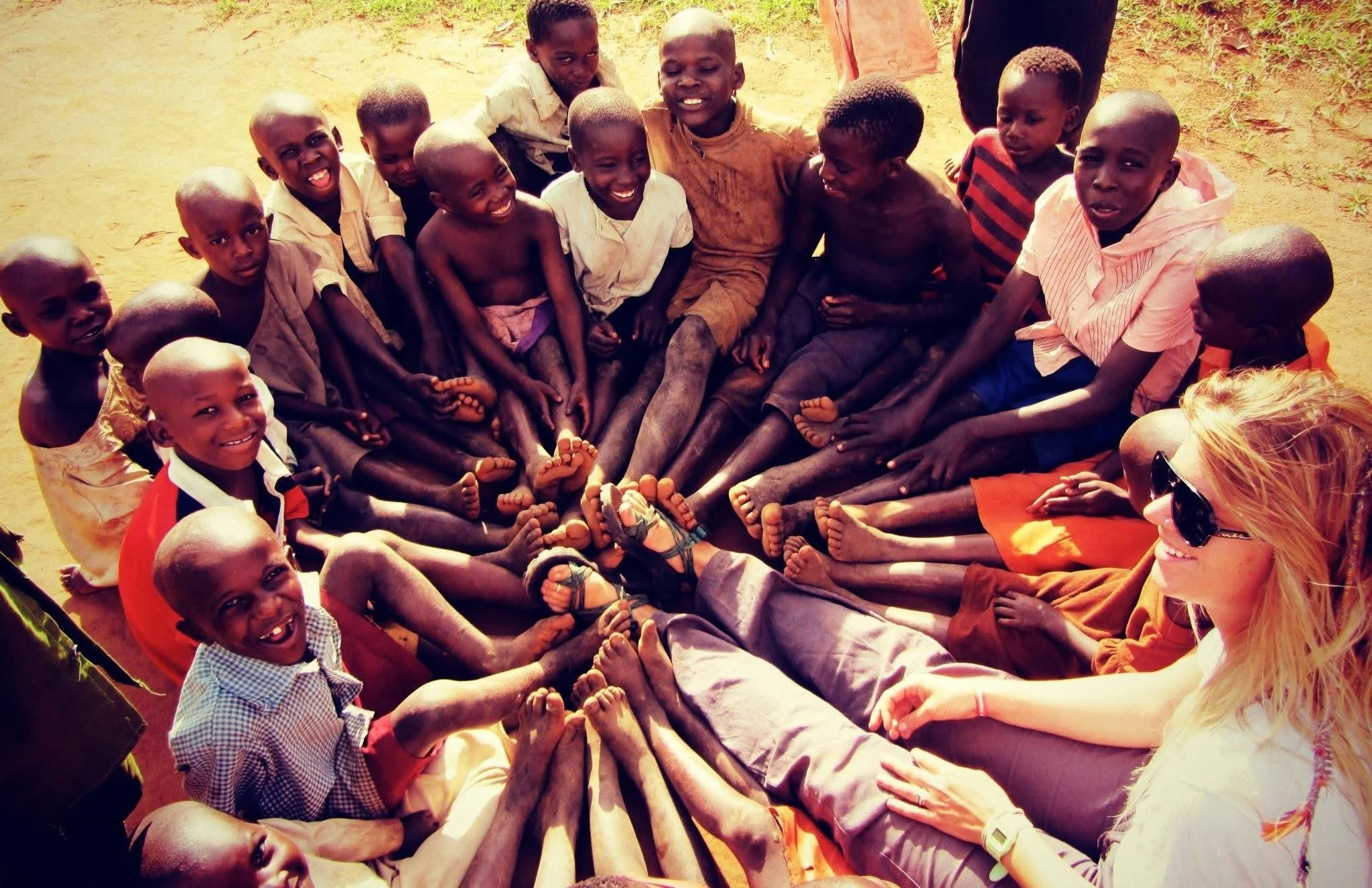 A group of children and a woman sitting in a circle with their legs extended inward, all touching at the center, on a dirt ground during daytime.