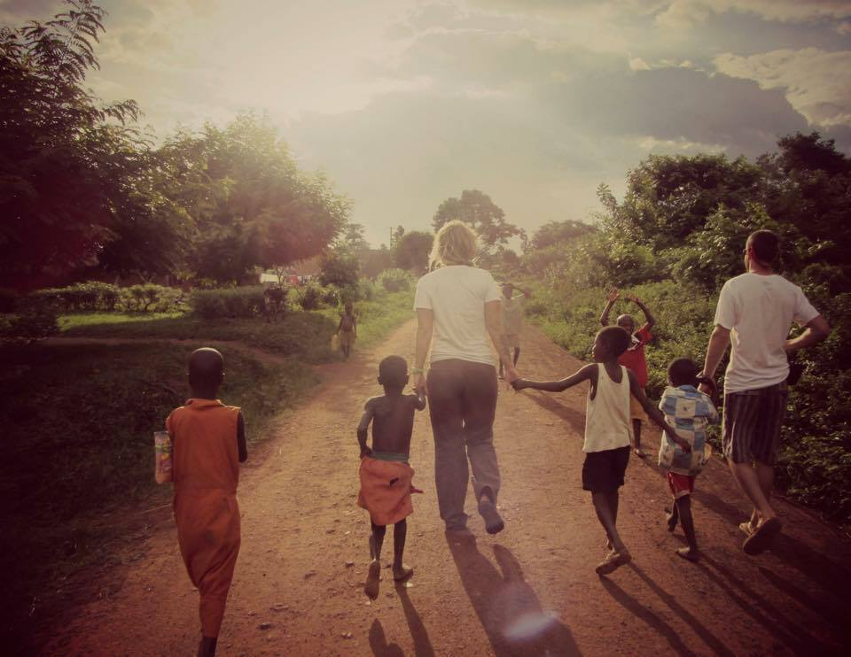 People walking down a dirt path in a rural area with children and adults, surrounded by greenery and trees under a partly cloudy sky.