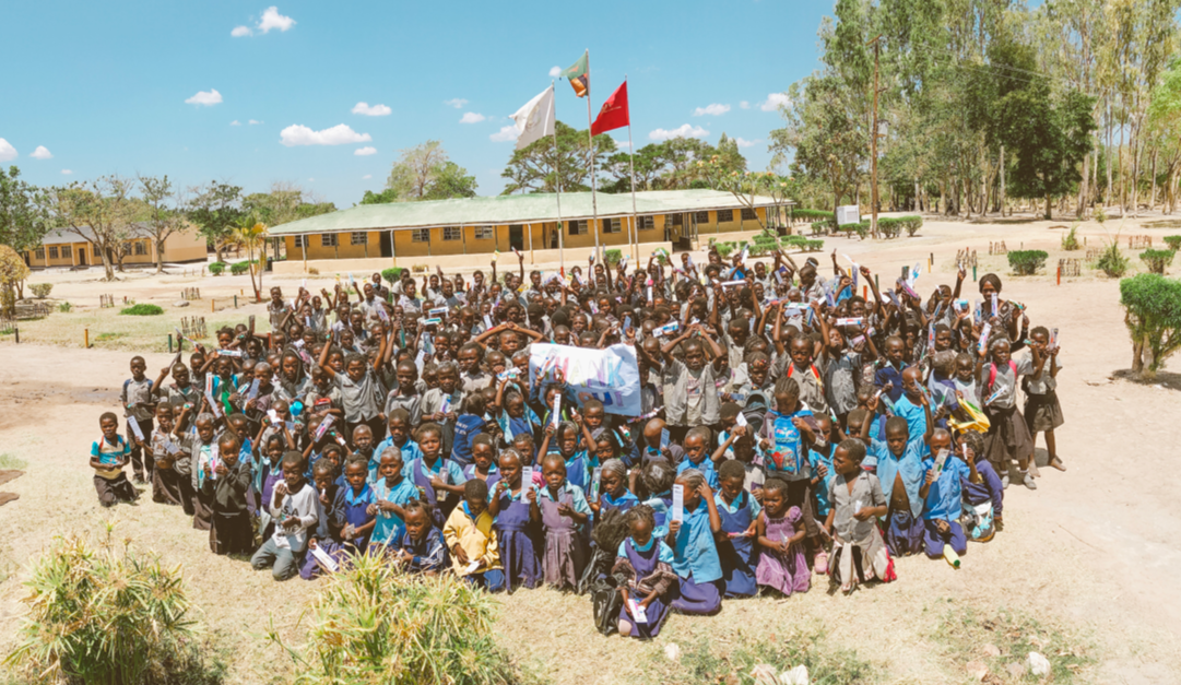 Large group of school children and teachers gathered outdoors on a school campus, holding signs and raising their hands in celebration.